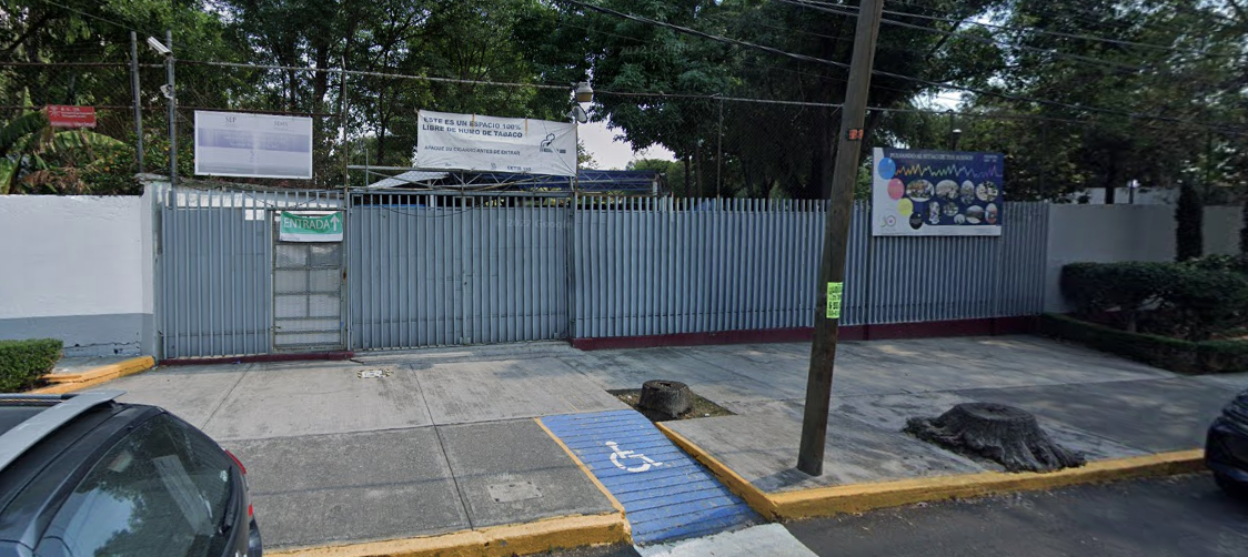 Fotografía en la que se muestra una explanada que tiene una rampa de acceso. Al fondo de la explanada está la entrada del plantel, conformada por una reja gris y muros blancos que rodean las instalaciones.